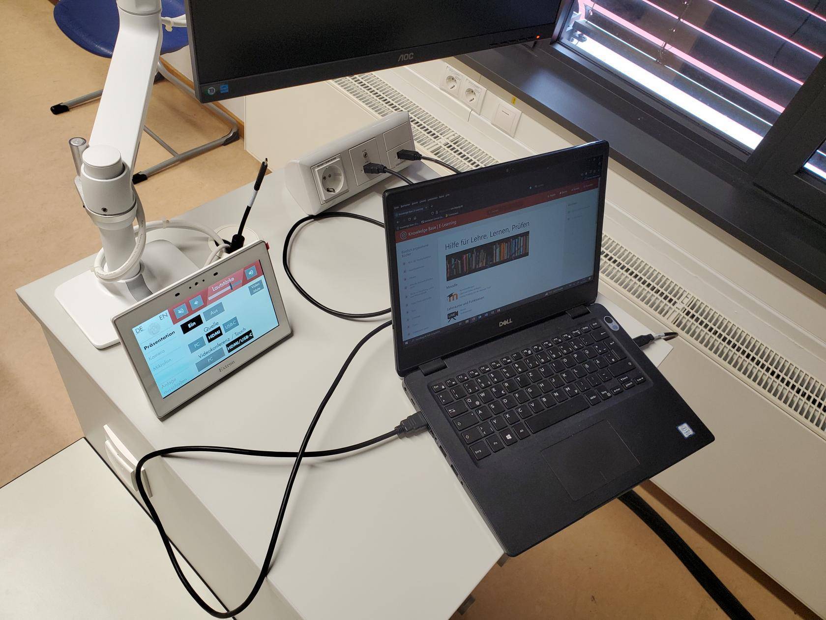 Dozierendenpult mit eingeschaltetem Mediensteuerungs-Touch-Display und Monitor auf Schwenkarm, darauf ein Laptop, der per HDMI- und 3.0-USB-B-Kabel mit dem Ansteckfeld verbunden ist. 