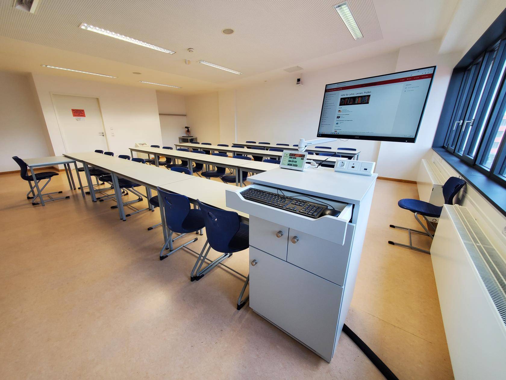 im Vordergrund eine Dozentenpult mit Mediensteuerung, PC und Monitor auf Schwenkarm, dahinter Tischreihen mit Stühlen, seitlich an der Wand eine kleine Garderobe, an der hinteren Wand eine Kamera