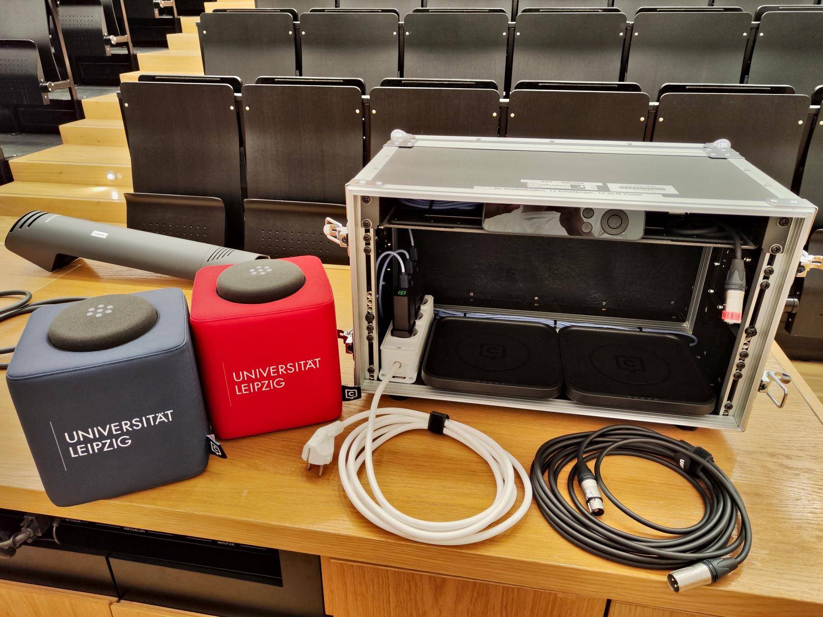 auf Dozentenpult stehen: graues und rotes Wurfmikrofon, geöffnete Transportbox mit Empfänger und Ladestationen, davor weißes Stromanschlusskabel und schwarzes Audiokabel