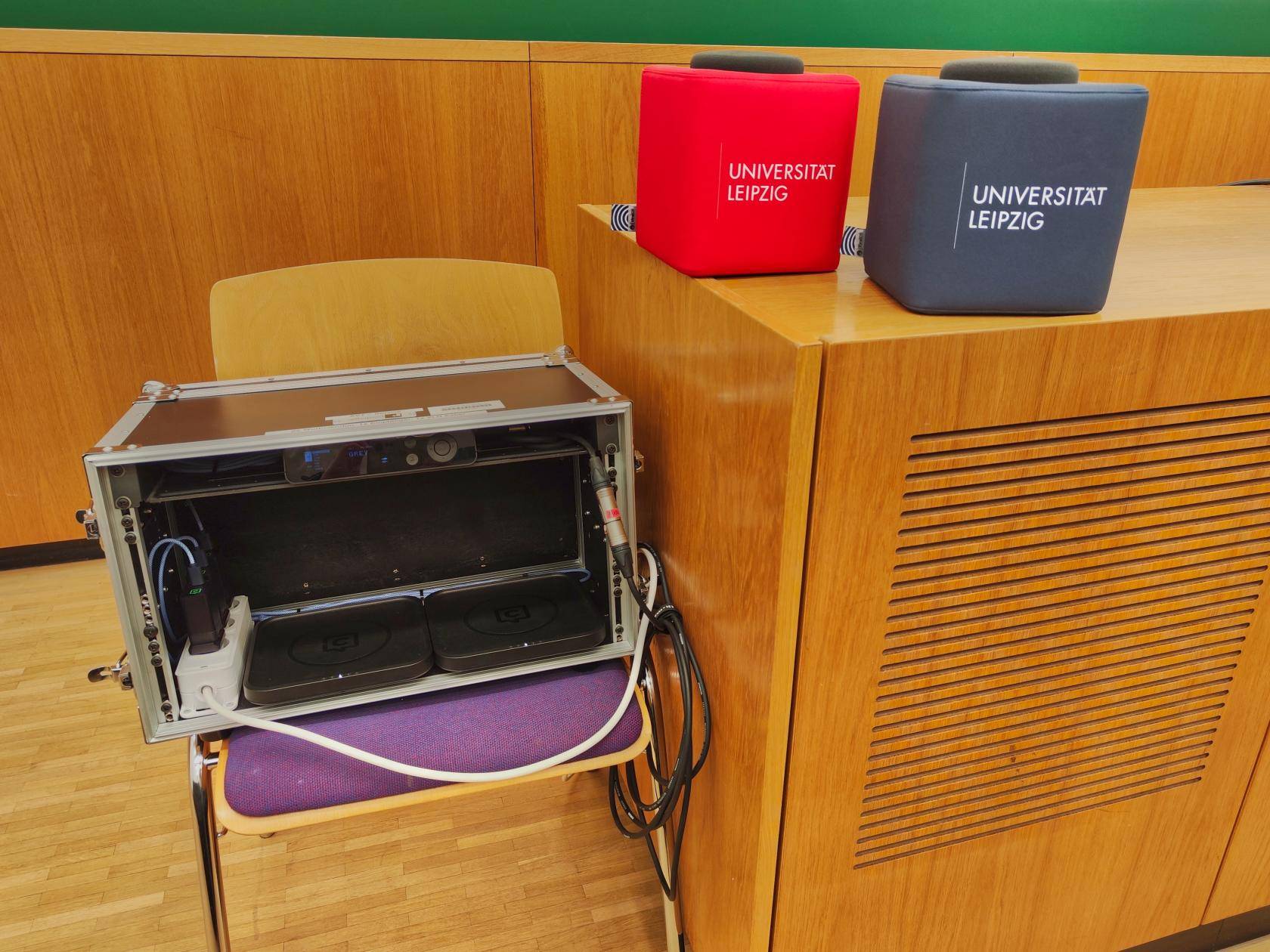 Stuhl neben Dozentenpult, auf dem Stuhl steht die geöffnete Transportbox, auf dem Dozentenpult stehen zwei Wurfmikrofone