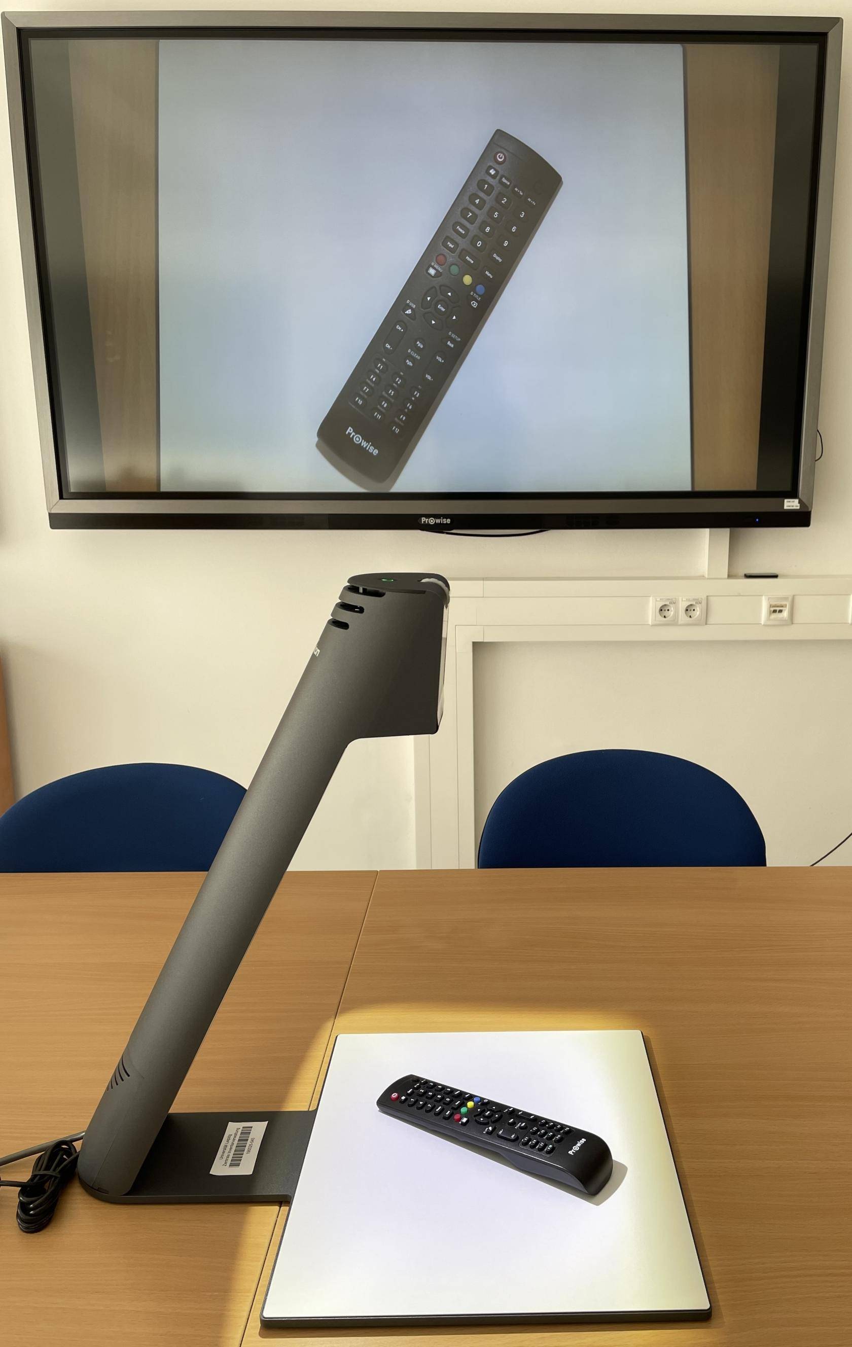 auf einem Tisch aufgebaute Dokumentenkamera, auf deren Vorlage eine Fernbedienung liegt. Im Hintergrund ein Bildschirm an der Wand, der das Bild der Fernbedienung zeigt